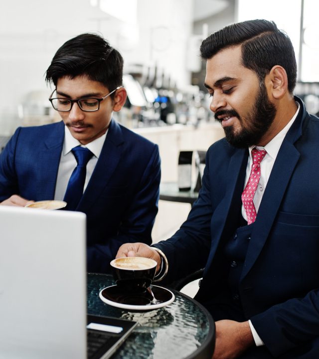 Two indian business man in suits sitting at office on cafe, looking at laptop and drinking coffee.